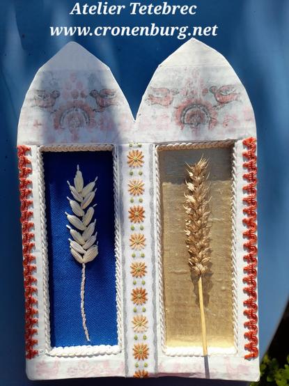 Showing the whole shrine. It is formed like a Gothic double window of a church. The main area consists of rectangular recesses with the ears of corn, blue on the left, and golden on the right. A machine-embroidered border with flowers runs lengthwise down the centre, and a brick-red braid runs along the sides. The shrine is painted with gesso. Under the gesso, traditional embroidery patterns in red and black become visible in the arches and at the lower edge. More on the details in the other photos.