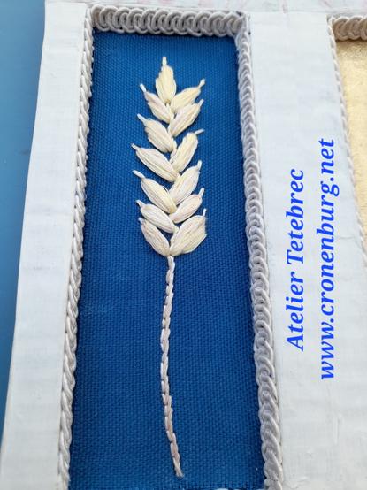An embroidered ear of corn on blue fabric in a rectangular recess of a cardboard shrine. The edges are lined with fine white braid, and the shrine is painted with white gesso.