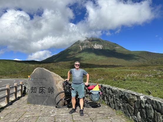 My sweaty self posing for a shot next to the Shiretoko Pass summit sign. It’s a big rock. Mount Rausu is in the background with some big puffy clouds.