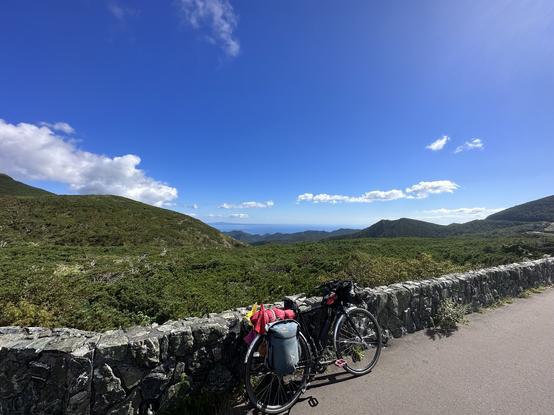 View down the descent to the south side of the peninsula from the summit. My bike is leaning against a rock wall and the hills are all lovely green.
