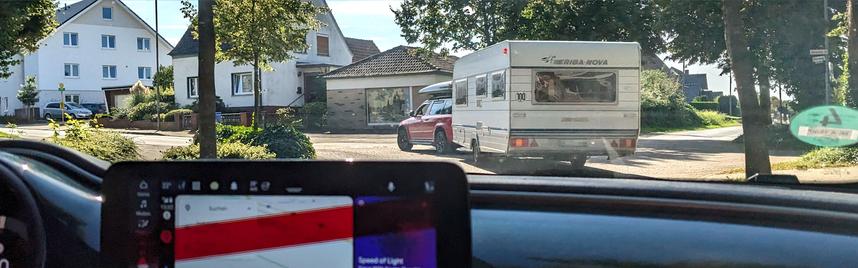 Ein Wohnwagen wird von einem PKW in einer Linkskurve gezogen. Das Bild ist aus einem dahinter fahrenden PKW aufgenommen und man sieht einen Teil des Armaturenbretts.