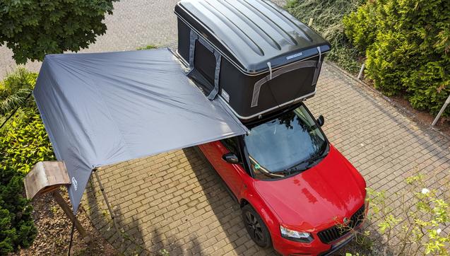 Ein PKW mit einem aufgeklappten Hartschalendachzelt auf dem Dach. Auf der Beifahrerseite ist eine Markise ausgefahren. Das Bild ist von schräg oben aufgenommen, so dass man auf das Dach des Dachzeltes schaut.