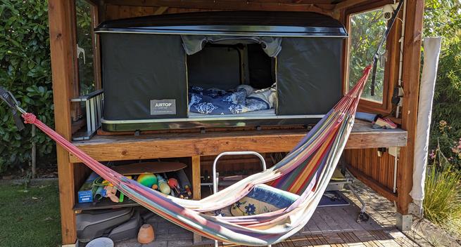 Ein aufgeklapptes Dachzelt mit Bettzeug steht  in einem Holzunterstand. Davor ist eine Hängematte gespannt.