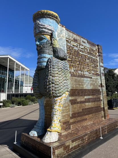 Photo of a Lamassu style statue by Michael ranowitz outside Towner gallery in Eastbourne, made from metal date boxes. Classic human head, assyrian style headpiece and beard, bull feat. Gold and light blue colours
