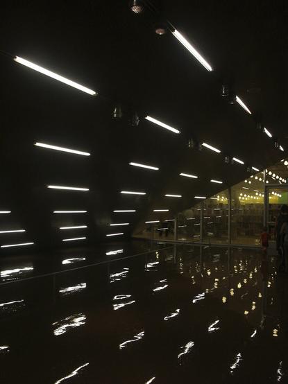 Bolts of light appear to fly through the dark space of a library, transforming into fire balls as they pass the glass wall into the main space.