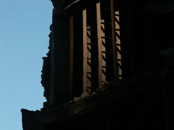 A clear blue sky is broken by the inky black darkness of a ruined building. In the middle of the darkness, light pools on rusty, tired beams…
