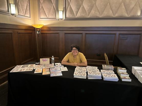person with curly hair in a yellow shirt sitting at a table and smiling, with stacks of the “bite size bash” zine on the right, one copy of every zine she’s published on the left, and a wooden box displaying zines