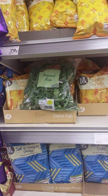 A supermarket shelf with a bag of kale discarded amongst bags of cheese puffs.