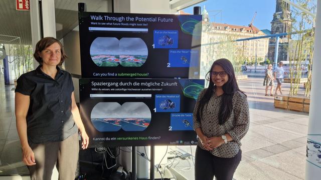 Foto im COSMO Wissenschaftsforum. Verena Maleska (li) und Tasnim Marzan Oyshi (re) stehen vor einem großen Bildschirm, auf dem gezeigt wird, was man in ihrer Virtual-Reality-Anwendung "FloodVis" sehen kann. Überschrift auf dem Monitor "Spaziergang durch die mögliche Zukunft".