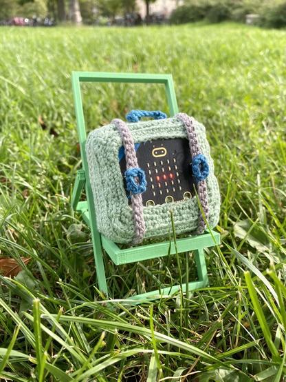 A pastel green crocheted travel suitcase with grey straps and blue handles. The front of the case is embedded with a Micro:Bit, which is an electronic microcontroller board with a grid of LEDs. Some of the LEDs are glowing and the straps cover the buttons on the board. The suitcase rests on a 3D printed deck chair laid on some grass in a park.