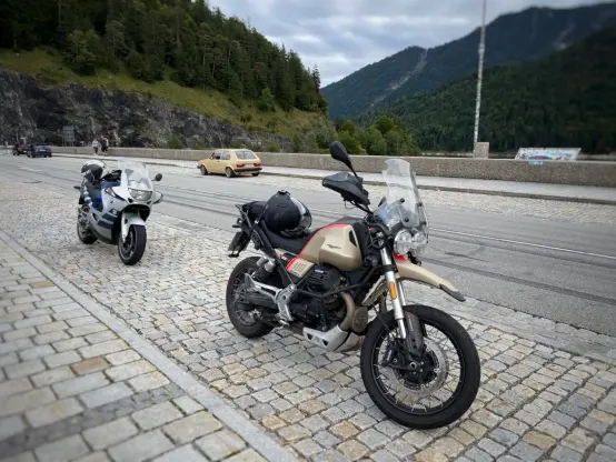 Zwei Motorräder und ein Auto stehen am Rande der Straße. Im Hintergrund sieht man Berge und ein Stück wolkigen Himmel.