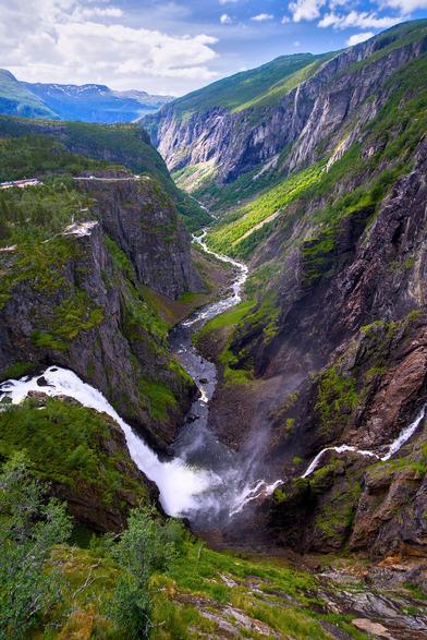 Ein Wasserfall im Vordergrund stürzt in ein Tal. Das Tal erstreckt sich zum oberen Bildende. Die Hänge des Tals sind grün vom Bewuchs und grau durch den Stein.