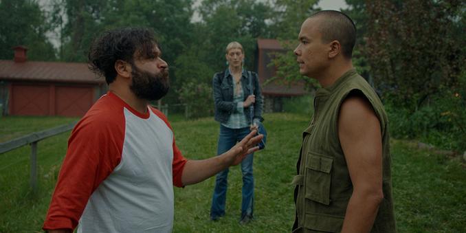 Three people stand in a back yard. Two men look like they are having a serious tension fraught discussion. A blond denim-clad woman looks on from a distance.