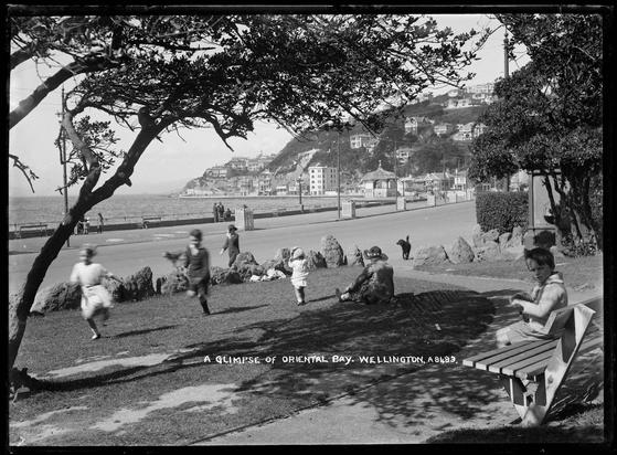 Black-and-white photo: Glimpse of Oriental Bay, Wellington. Showing a view of Oriental Bay, Wellington, with children playing in park area, woman sitting on grass; child on park bench (right); rotunda (right rear); buildings on hill and along Oriental Parade; people on footpath. 1910s–1923. Photographer, Frederick George Radcliffe (1863–1923). Auckland Libraries Heritage Collections 35-R1779. https://kura.aucklandlibraries.govt.nz/digital/collection/photos/id/274848