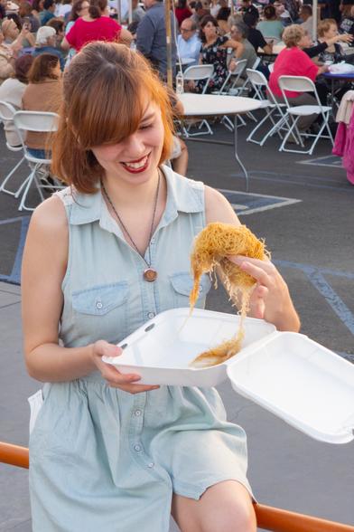 Photograph of Lety leaning against the railing at the Saint Nectarios National Shrine. She's wearing a light blue denim button-up dress and holding very soggy kataifi above a takeout tray. Beneath the event tent behind Lety are hundreds of people in nice, comfortable folding chairs.