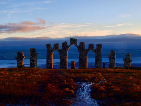 A photo of the Fyrish Monument  3 crenellated stone arches with 2 smaller graduated columns at either end and a small round post in the centre arch. A folly of sorts. An elevated view of the Cromarty Firth behind the monument. ice blue water darkened by the fading light. A pink tinged sky above and in the foreground frozen heather tinged russet with the glow of sunlight.
