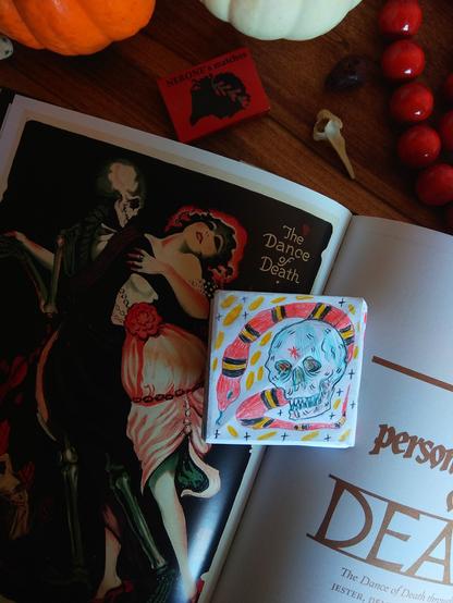 A little piece of paper, the artist has drawn a skull and a snake on it. The snake is red with black and yellow straps. On the background: a book with a spooky art (woman dancing with a skeleton), some pumpkins, a matchbox (red), a bone, red jewelry.