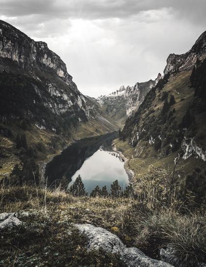 A deep valley, at the bottom of which lies a lake reflecting steep mountains and the cloudy sky. The sun is peeking gently through the cloud cover.