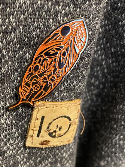 An orange metal and enamel pin shaped like a feather and a cork 10tree patch on a grey fabric. The pin has imagery of a “boy and girl, the moccasins that walked them home, the sun that guided them, and their wings that allow them to visit us here, in the earth world”