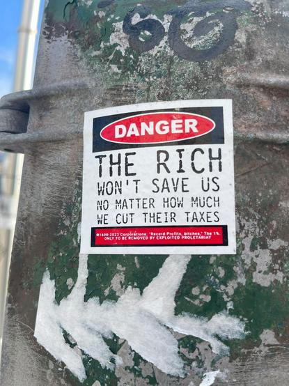Danger sticker on a pole that says "The rich won't save us, no matter how much we cut their taxes"