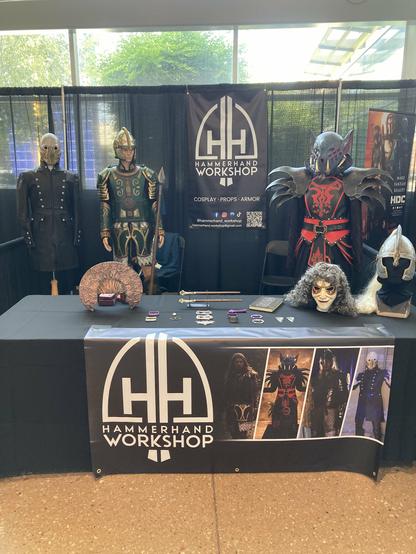 Photo of a booth at Omaha Comic Con, displaying my banners, threemannequins wearing my costumes, and several assorted helmets and masks on the table.