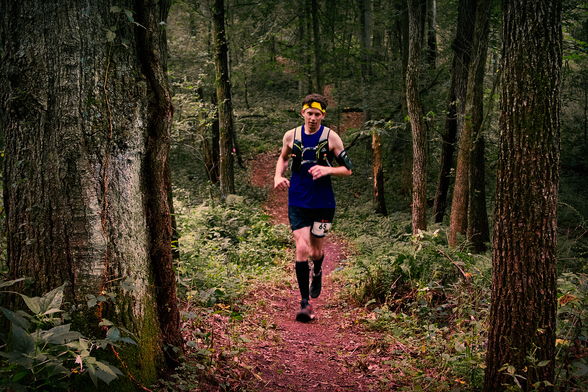 Photo of me running through a midwestern forest during a 50K race. I am wearing black calf sleeves, black shorts, a blue tank top, yellow headband, and a hydration vest.