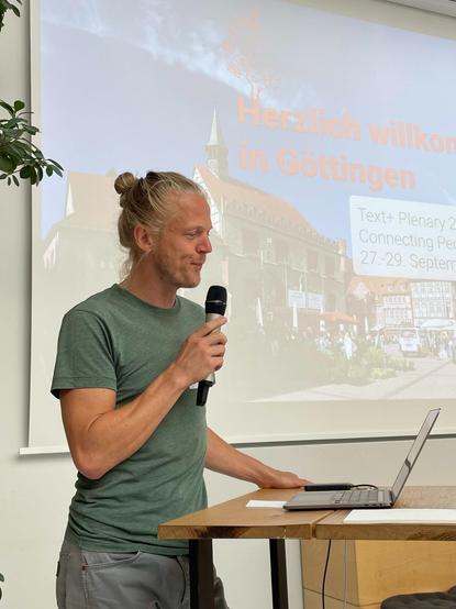 Mann mit Dutt spricht ins Mikrofon. Er steht vor einem Laptop. Im Hintergrund ist eine Präsentation auf einer Leinwand zu sehen mit dem Text: Herzlich willkommen in Göttingen. Text+ Plenary 2023.