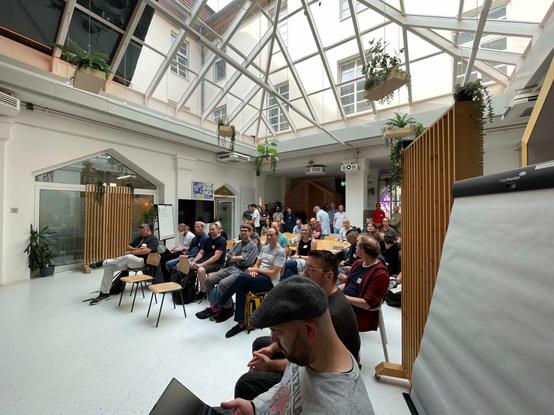 Weitwinkelfoto von einer Gruppe, die auf Stühlen in einem Raum sitzt, einige haben einen Laptop auf dem Schoß. Der Raum hat ein Glasdach und es hängen viele grüne Pflanzen vom Dach.
