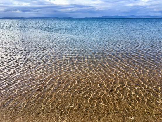 photo of a crystal clear sea with piles from a breeze. Sand in the foreground and shades of blue into the distance with a strip of blues hills distant and a cloudy sky