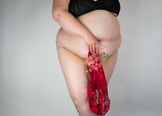 Lindley's belly, thighs, arms and chest are shown against a plain background. She's wearing a black bra, is holding a pair of red panties over her pubic area, and has pale skin and stretch marks.