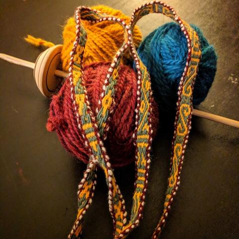 Three large balls of wool yarn (in mustard yellow, blue, and red). Between and the yarn is a single empty wooden spindle. On top, two patterned woven narrow bands -- each is dark red on the outer edges, green and yellow figures running down the centers, and white cotton polka dots on the outside edges (similar to white beads appearing on the edges)