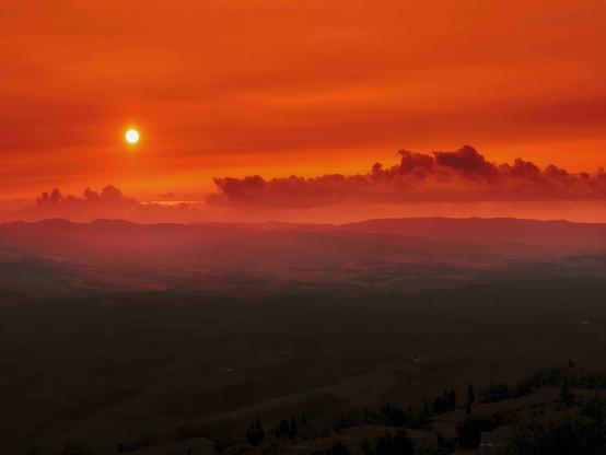 A wide view over a Tuscan landscape at sunset. From an elevated position you can vaguely see hills stretching towards the horizon, individual cypress trees, villages and houses. The sun turns the horizon a red-orange color and distant cloud formations are visible in the sky.