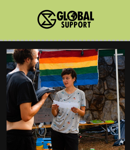 An XR volunteer organizer holding a sheet of paper at the Adriatic Climate Camp in Krk, Croatia. In the background is a multi coloured flag mounted on a stone wall. Another XR camp attendee stands to the left.