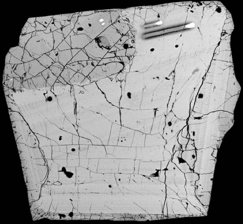 A backscattered electron image of a zircon crystal with multiple zones. The change in brightness of the zones are caused by variations in trace elements such as hafnium, titanium, and uranium. Two laser ablation trenches are present in the upper right corner, where material was sampled for analysis.
