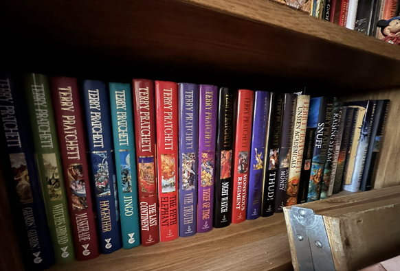 A wooden shelf full of hardback Terry Pratchett books, including classics like Hogfather and Monstrous Regiment