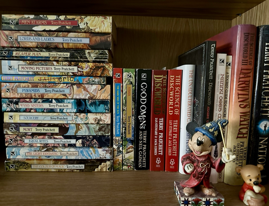 A shelf full of Terry Pratchett Discworld and Good Omens books - accompanied by a small figurine of Sorceror Mickey Mouse