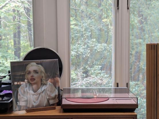 Glüme - Main Character LP.

Glume, highly made up, looking a bit like Marilyn Monroe, in a silk top.

The pink LP plays on a vintage Bang & Olufsen Beogram 4000 linear-tracking turntable to the right. The dustcover is open, and the album, tonearm and surround are reflected in its underside.