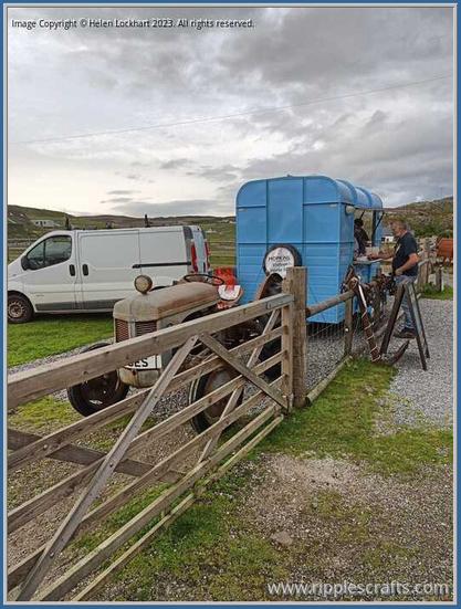 Photo shows a gate, behind which is a very old tractor and a blue van in which folk are cooking fish and chips.  There is a modern white van alongside.  The sky is a mix of clear skies and cloud.