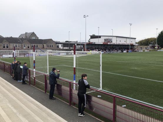 A handful of spectators at a football ground with concrete terracing, a primary school and low stand.
