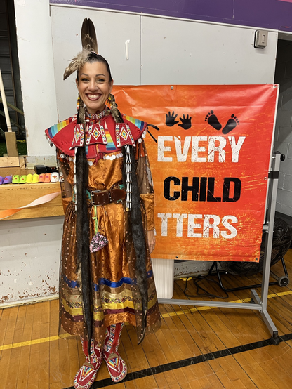 Mariah Dumont from Cold Lake First Nation, in an ornate orange ribbon skirt, her hair in elaborate braids, stands next to an orange sign that reads Every Child Matters.