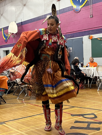 Mariah performs a fancy shawl dance, spinning and leaping in an elaborately embroidered orange shawl and ribbon skirt.