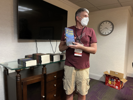 Joshua Palmatier (a man in burgundy shirt and khaki shirts and a white face mask) holds a copy of SOLAR FLARE. A TV screen and clock on the wall behind him.