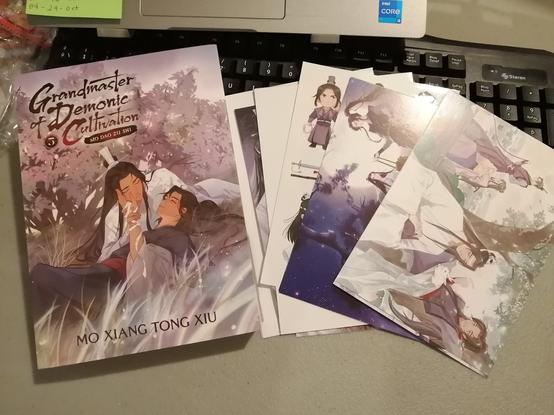 Volume 5 of the physical copy of Grandmaster of demonic cultivation next to some postcards and a sticker sheet. They're on a desk, over a black keyboard.