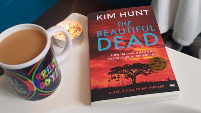 Book The Beautiful Dead by Kim Hunt next to a mug of tea with a Rebel Dykes logo on it next to a candle, all on the side of a bath tub.