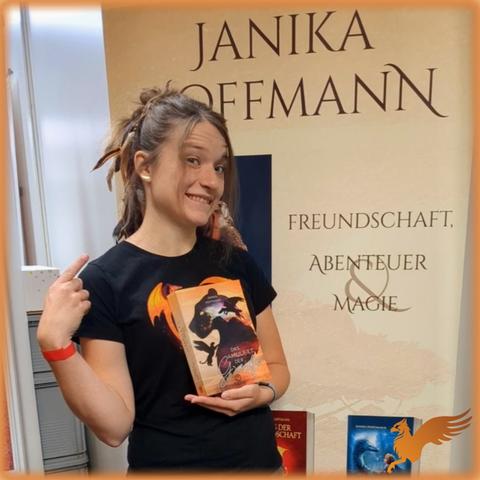 Frau Ende 20 steht vor einem Roll-Up mit Beschriftung "Janika Hoffmann: Freundschaft, Abenteuer & Magie". Sie hält ein Buch vor der Brust, auf dem Greife abgebildet sind.
In die langen Haare sind rotbraune Federn eingeflochten.