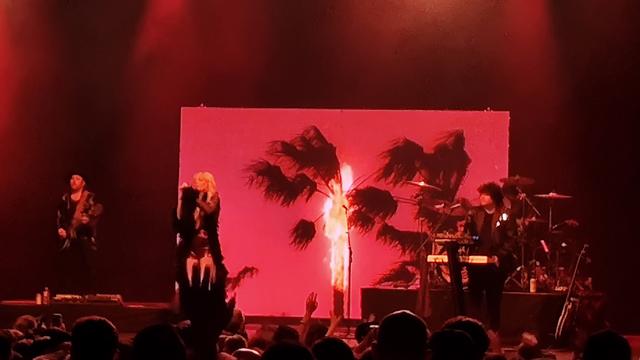 The band, Cannons, performs live before an audience in Fox Theater Oakland for the final show of their first headlining tour: Heartbeat Highway. Red lighting and a screen displaying palm trees against a red sky with one tree in the middle on fire sets the mood.