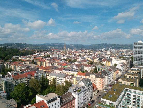 Blick von oben auf die Stadt bei Sonnenschein. Im Vordergrund viele Hausdächer. Im Hintergrund Berge und dazwischen der Dom.