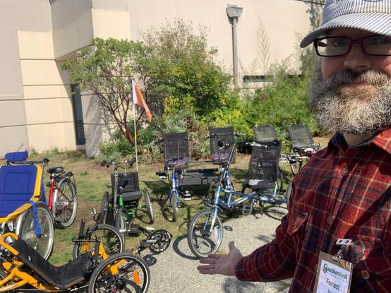 A guy with glasses, beard, and a hat standing in front of a variety of adaptive bicycles.
