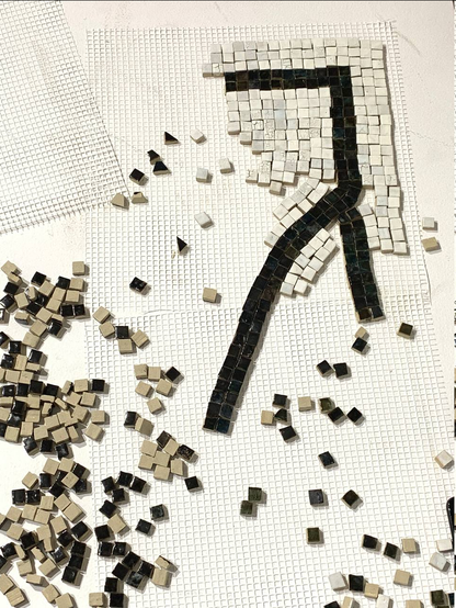 A section of mosaic tiling in progress over fiberglass mesh, surrounded by loosely scattered tesserae. The tiling shows a thick black line branching out into two lines, enveloped by off-white tesserae with some crackle effects.