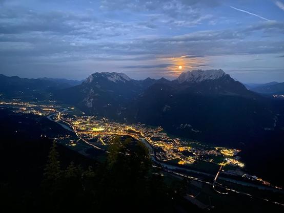 Abendaufnahme von einem Gipfel ins Inntal. Zu sehen ist der Fluss im Tal und die vielen Lichter der Menschen und entlang der Straße, auch die gegenüberliegende Bergkette mit felsigen Gipfeln. Es ist fast schon dunkel, hinter den Bergen geht gerade die orange glühende Sonne unter und färbt die Wolken ums ich her.